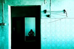 Blue Room - figure sits hunched in the distance of a doorway in a blue-tinged tiled room in a state of disrepair.