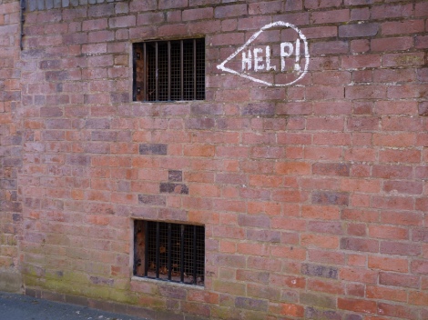 A brick wall with two barred windows near the top and bottom of the image. Next to the top one, a graffiti bubble says: "Help!"
