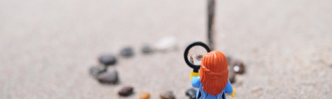 A lego figurine with long red hair stands on a sand beach in front of a circle of small stones, which they inspect with a magnifying glass.
